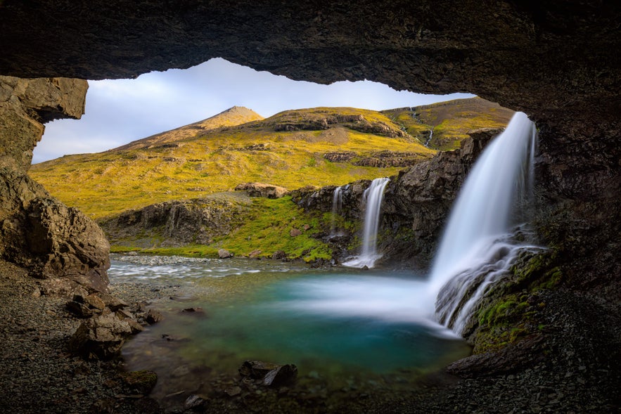 Skutafoss Waterfall flows into a clear pool, as seen from inside a cave opening, with grassy hills rising in the background.