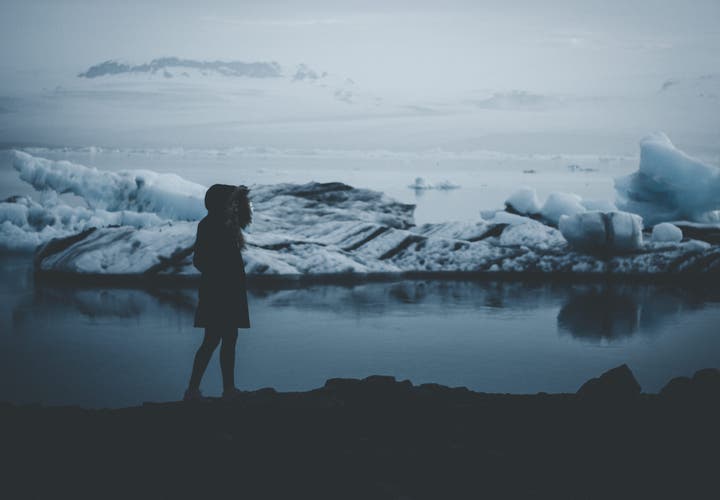 Silhouetted figure in a hooded coat standing by an icy lagoon in Iceland, surrounded by floating icebergs and a misty, moody landscape.