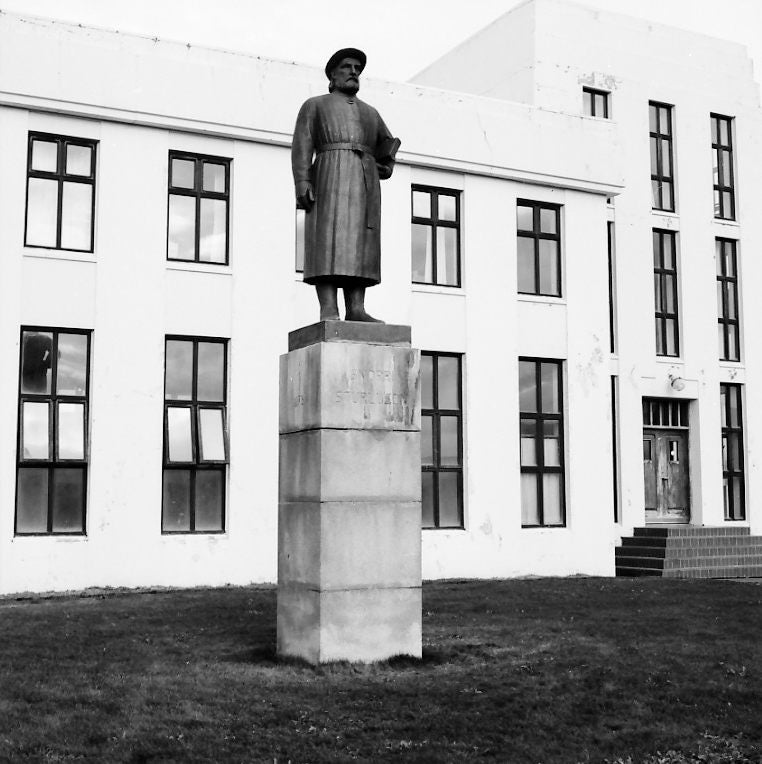 Black-and-white photo of a statue of Snorri Sturluson standing on a tall stone pedestal in front of a modernist building with large rectangular windows in Iceland.