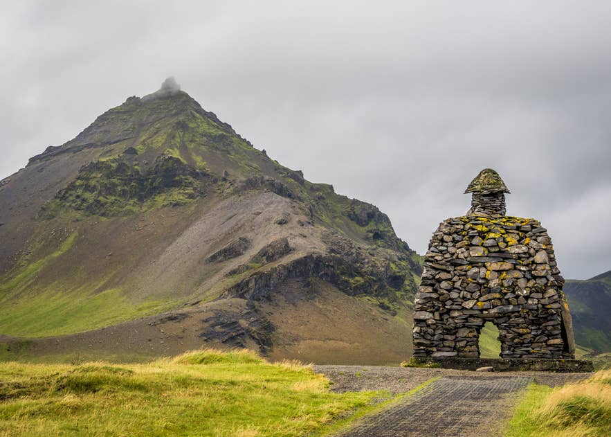 Eine steinerne Statue von Bárður Snæfellsás vor einem dramatischen, moosbedeckten Berg auf der Halbinsel Snaefellsnes in Island, aufgenommen bei bewölktem Himmel.