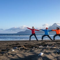 Reisende som praktiserer mindfulness-yoga på en svart sandstrand i Østfjordene med snødekte fjell i bakgrunnen.