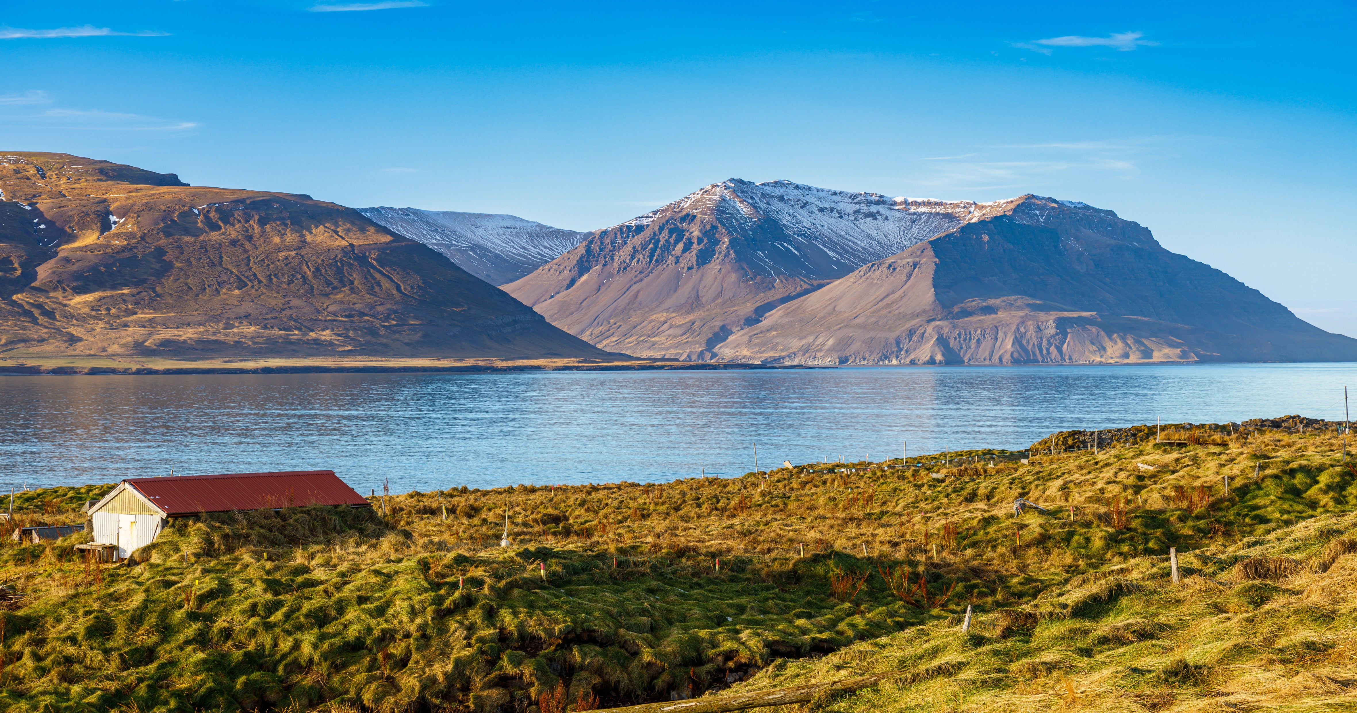 Scenic view of Borgarfjordur fjord in West Iceland, with snow-dusted mountains reflecting in calm blue water and a red-roofed farmhouse in the grassy foreground.