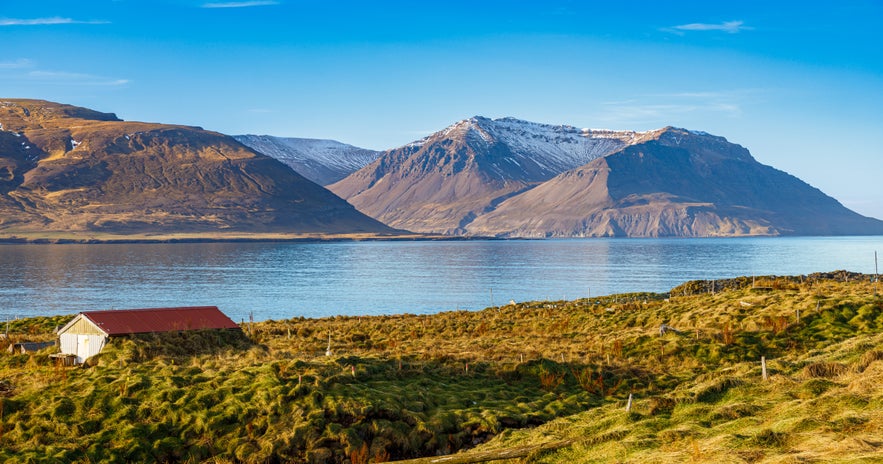 Scenic view of Borgarfjordur fjord in West Iceland, with snow-dusted mountains reflecting in calm blue water and a red-roofed farmhouse in the grassy foreground.