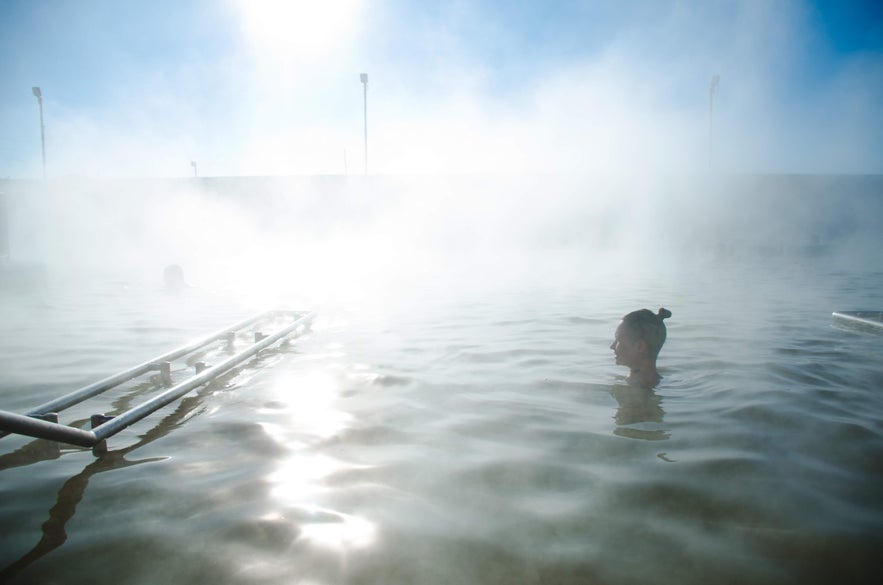 A person enjoys a steaming geothermal pool in one of Reykjavik's top spas, with a ladder and bright sky.