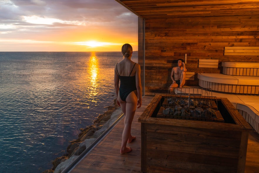 A person in a swimsuit stands on a deck at Sky Lagoon Spa, admiring the sunset from a sauna area.