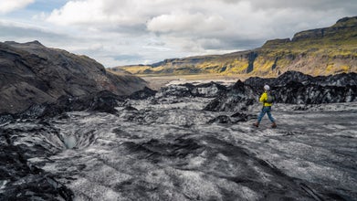 A solo hiker in yellow explores the rugged ice fields of Solheimajokull Glacier with mountains in the distance.