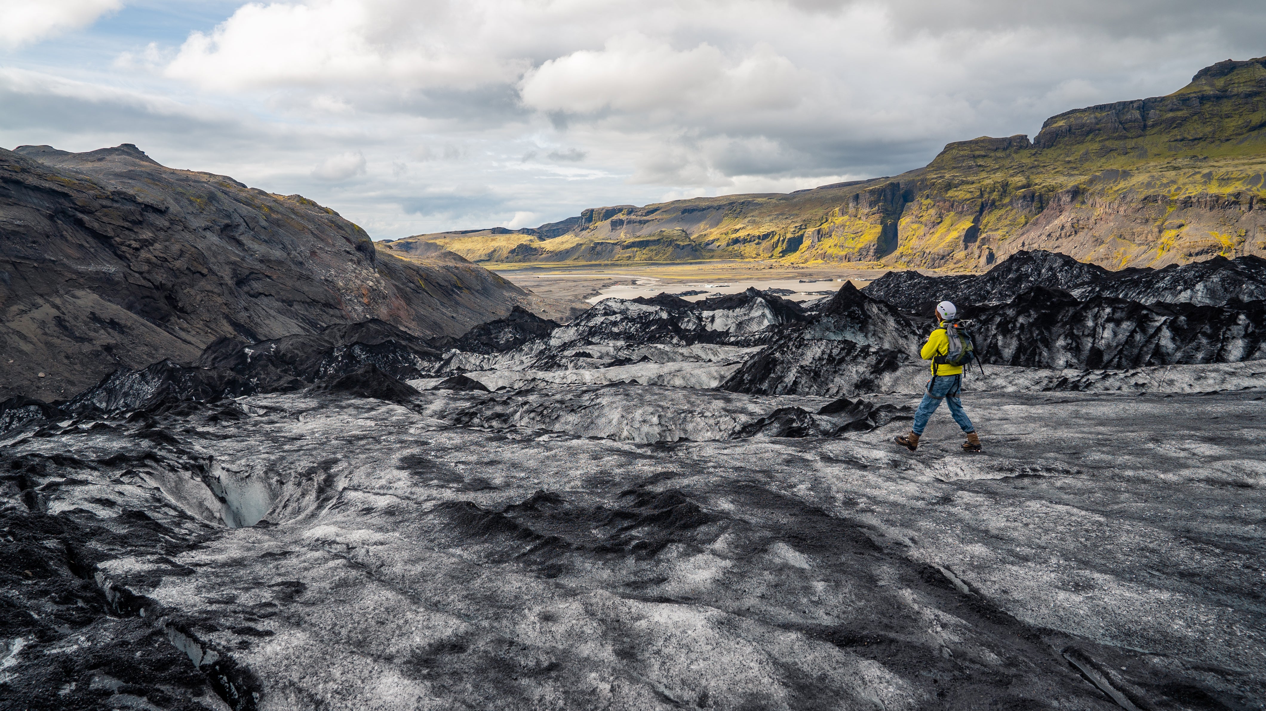 A solo hiker in yellow explores the rugged ice fields of Solheimajokull Glacier with mountains in the distance.