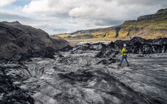 A solo hiker in yellow explores the rugged ice fields of Solheimajokull Glacier with mountains in the distance.