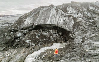 Person with backpack near the outlet river of a glacier, standing in front of dark glacial ice and meltwater flow.