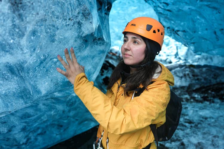 A woman gently touches the smooth, frozen wall of a glowing blue ice cave in Iceland.