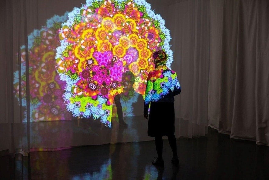 A person stands in front of a vibrant, colorful digital projection resembling a kaleidoscope of flowers and geometric patterns in an art installation at the House of Collections in Reykjavík, Iceland.