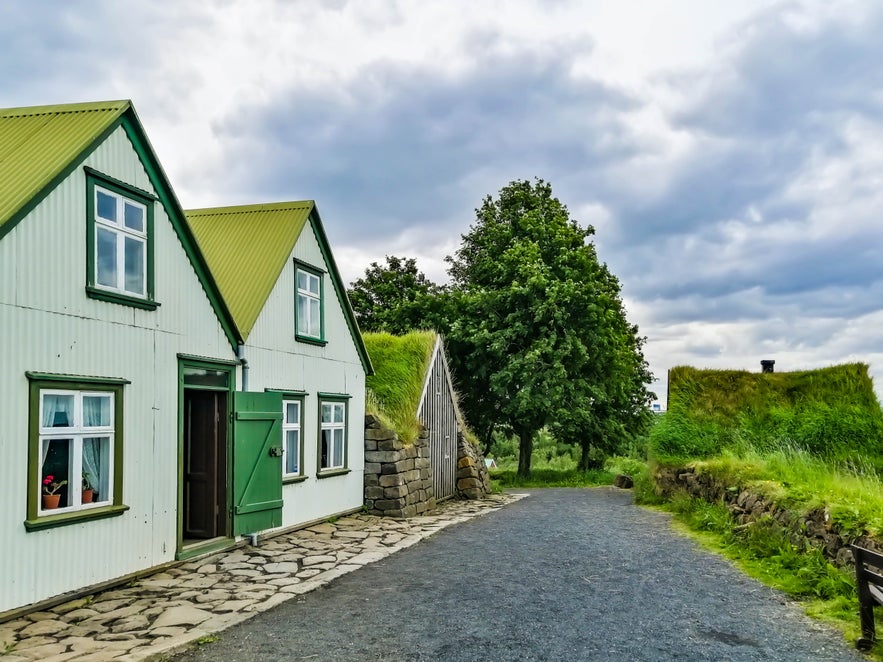 Traditional turf-roofed buildings and green-trimmed timber house at Árbær Open Air Museum in Reykjavík, along a gravel path lined with grass and trees.