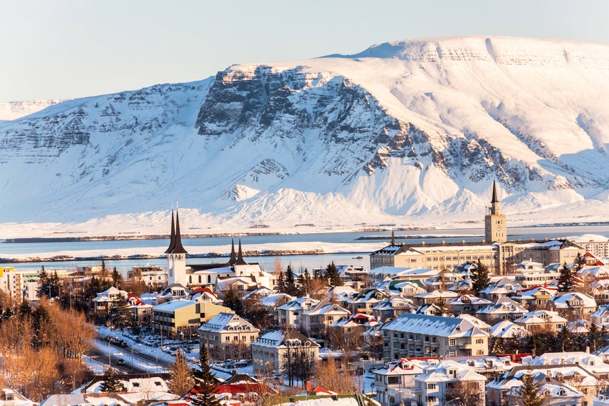 Snow-covered Reykjavík with colorful rooftops and church spires set against the dramatic backdrop of Mount Esja in winter sunlight.