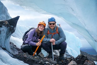 A couple wearing helmets and holding axes smile as they pose with a stunning glacial landscape behind them.