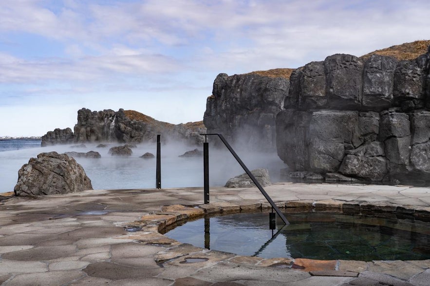 Steam rises from a geothermal infinity pool at Sky Lagoon, surrounded by volcanic rocks and overlooking the ocean.
