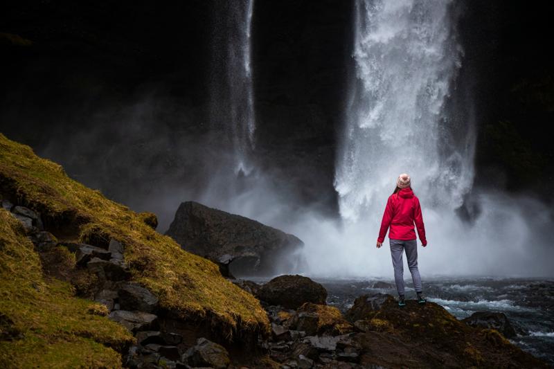 A person in a red jacket stands on rocks near the base of Kvernufoss waterfall, surrounded by mist and mossy cliffs.