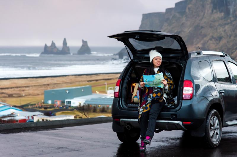 A traveler holds a map while sitting in the back of a car near Vik, Iceland, with Reynisdrangar sea stacks visible in the background.