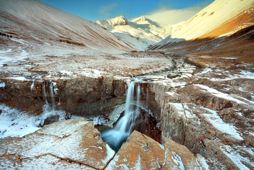 Skutafoss waterfall flows over a rocky cliff into a narrow canyon, framed by snow-dusted slopes and distant mountains in East Iceland.