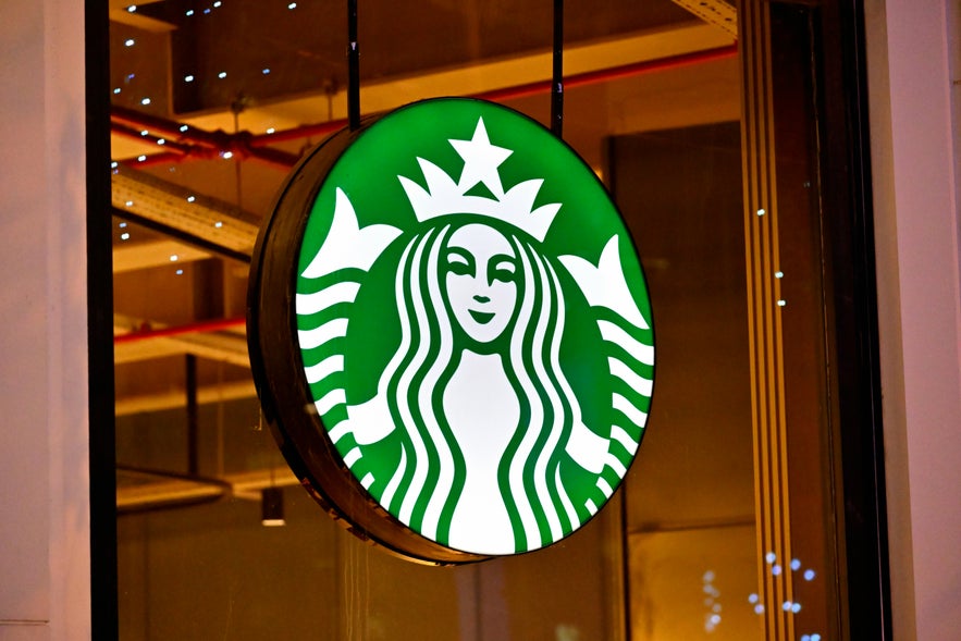Close-up of an illuminated round Starbucks sign featuring the iconic green and white siren logo, hanging outside a store with warm interior lighting and visible ceiling fixtures.