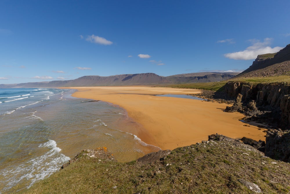 Wide view of Rauðisandur Beach with golden sands, clear waves, and cliffs under a blue sky in Iceland