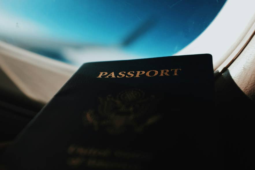 A dark passport resting on an airplane tray table beside a window, representing travel to Iceland