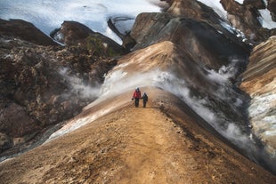 Two people walk through the Hveradalir geothermal valley in the Icelandic Highlands, with steam rising around the peaks and snowy terrain stretched out behind.