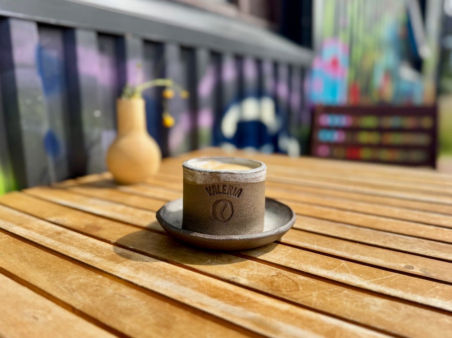 Cappuccino in a ceramic cup with the Valeria logo, sitting on a wooden table outside the café.