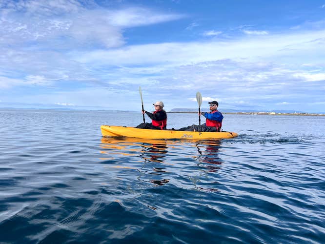 Two people kayaking a yellow tandem kayak on calm waters near Stykkisholmur on the Snaefellsnes Peninsula, under a bright blue sky with scattered clouds.