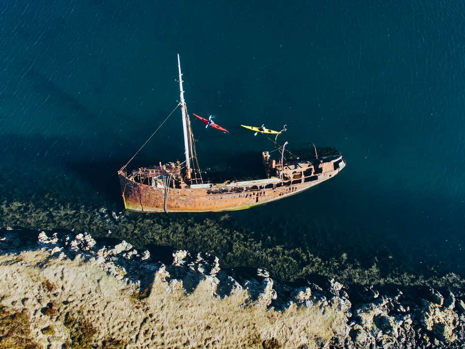 ภาพถ่ายทางอากาศของนักพายเรือคายัคสองคนกำลังพายเรือข้างซากเรือสนิมขนาดใหญ่ในน้ำตื้นใกล้ชายฝั่งที่มีโขดหินในเมือง Stykkisholmur บนคาบสมุทร Snaefellsnes