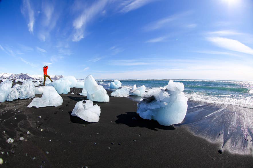 Un touriste en veste orange se tient sur les icebergs à la Plage de diamants en Islande, sous un ciel bleu parfaitement dégagé.