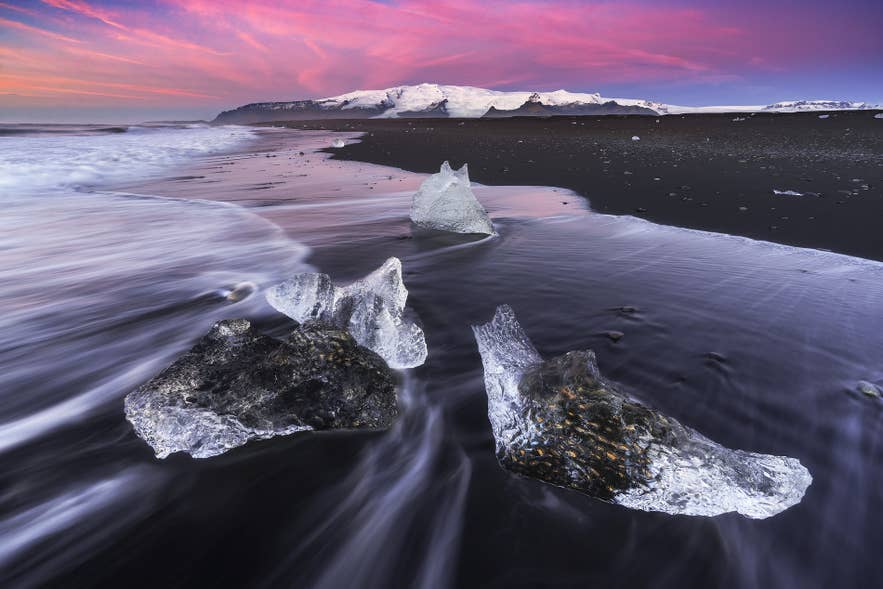 Des morceaux de glace glaciaire sont éparpillés sur le sable noir de la Plage de diamants sous un ciel rose.