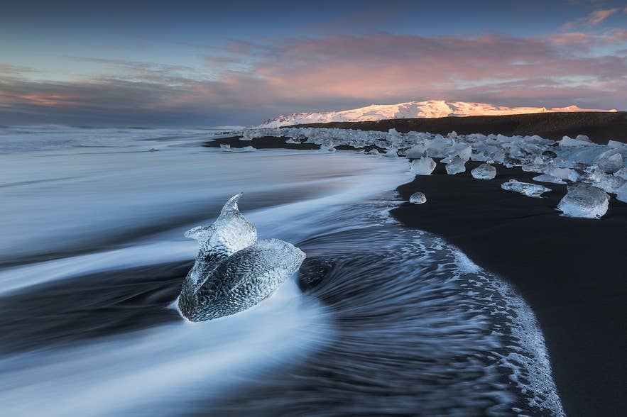 La Plage de diamants offre du sable noir, des morceaux de glace brillants et un glacier au loin sous un ciel dégagé.