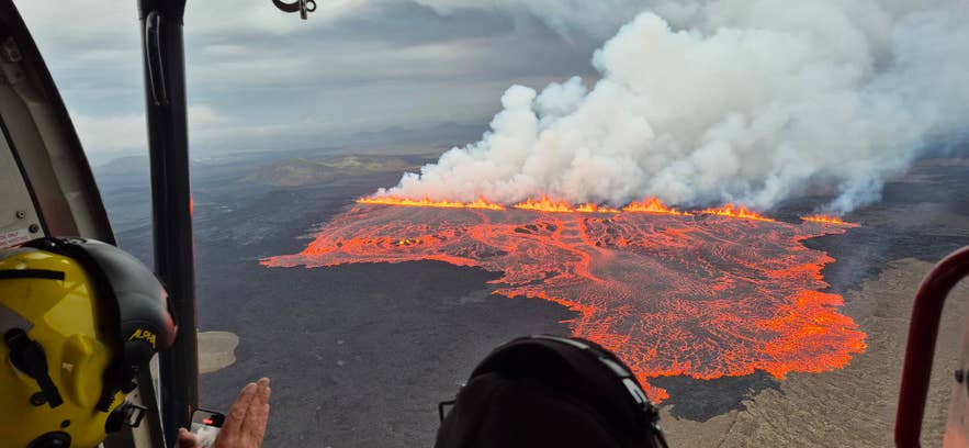 Lava flows from a volcanic fissure in Iceland as seen from a helicopter, with thick smoke rising into the cloudy sky.