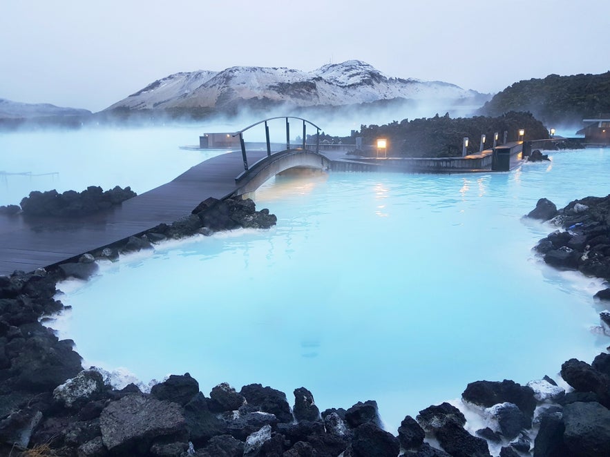 The Blue Lagoon features milky blue geothermal waters, with black lava rocks and steam rising into the sky.