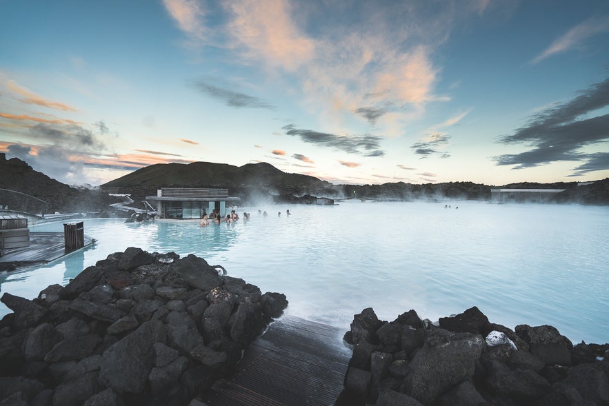 Visitors are enjoying a soak in the warm, milky blue geothermal waters of the Blue Lagoon in Iceland, surrounded by steam and lava rocks.