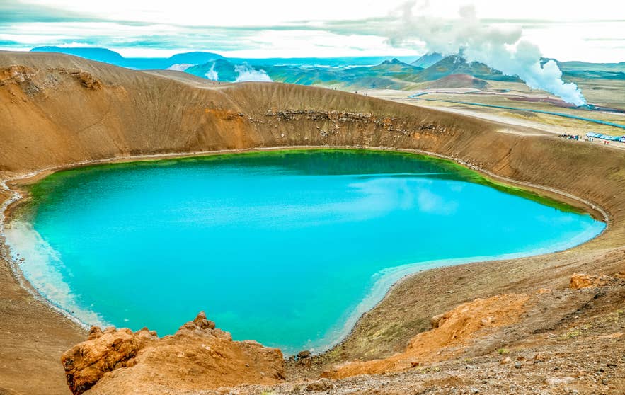 El brillante lago del cráter Viti en el norte de Islandia en julio, rodeado de laderas volcánicas rojizas y vapor geotermal a lo lejos.