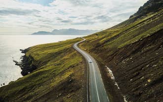 Car on a scenic coastal road, between lush mountains and ocean views on a summer self-drive tour in Iceland.