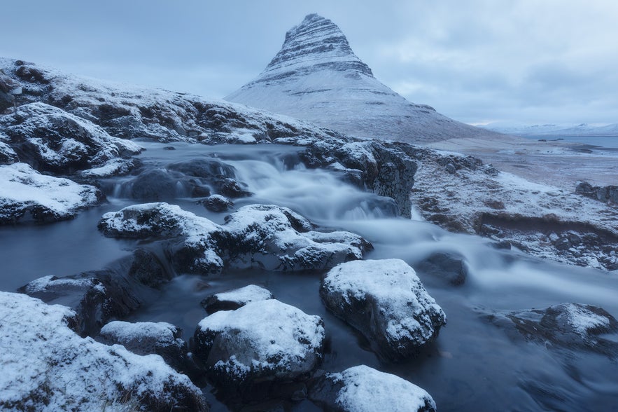 Kirkjufell Mountain in winter is seen with icy stones in the foreground.