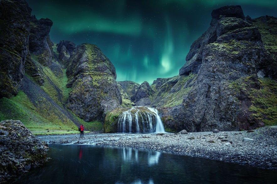 Northern lights dancing over Stjornarfoss Watefall near Kirkjubaejarklaustur with photographer in red