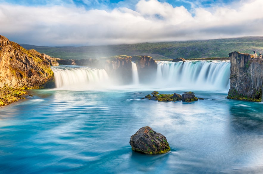 Water rushes over the wide cliffs of Godafoss Waterfall in Iceland, surrounded by lush green scenery on a summer day.