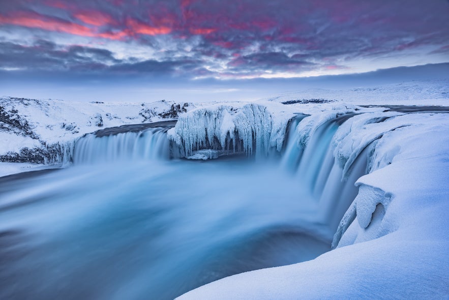 Water cascades down the icy cliffs of Godafoss Waterfall, with snow and ice covering the winter landscape in Iceland.
