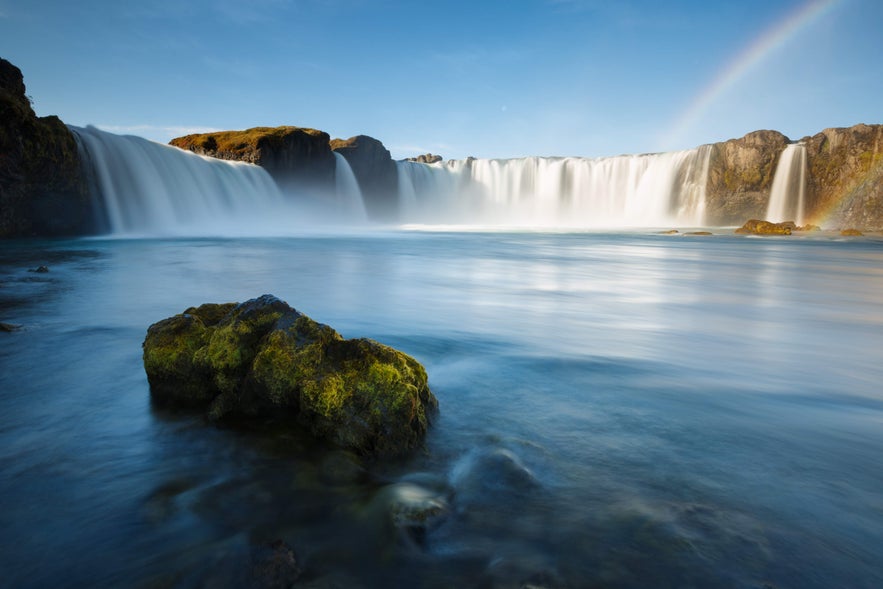 A rainbow forms over Godafoss Waterfall in Iceland as it rushes down rocky cliffs on a summer day.