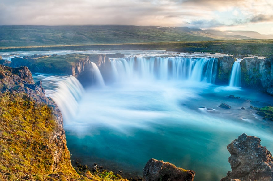 A picturesque view of Godafoss Waterfall in northern Iceland shows water cascading down mossy cliffs on a clear summer day.