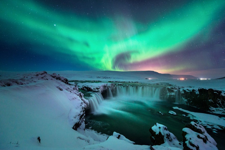 Northern lights glow in the night sky above Godafoss Waterfall, with water cascading over snow-covered rocks into the river below.