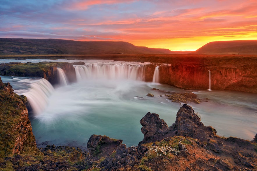 The sun sets behind Godafoss Waterfall, painting the Icelandic sky in warm colors as the water flows through the rugged landscape.