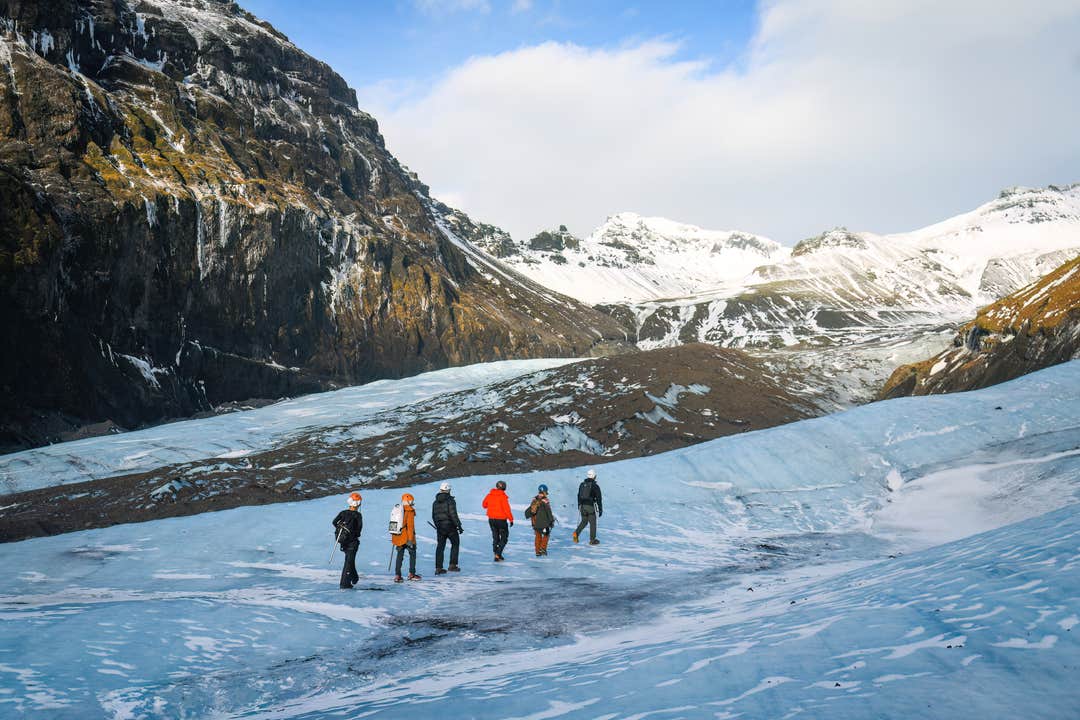Escursionisti attraversano il ghiacciaio Solheimajokull dal colore blu intenso in Islanda, circondati da picchi innevati e cielo nuvoloso.