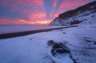 Snow-covered Reynisfjara Black Sand Beach in Iceland at sunset, with dramatic crepuscular rays lighting the sky.