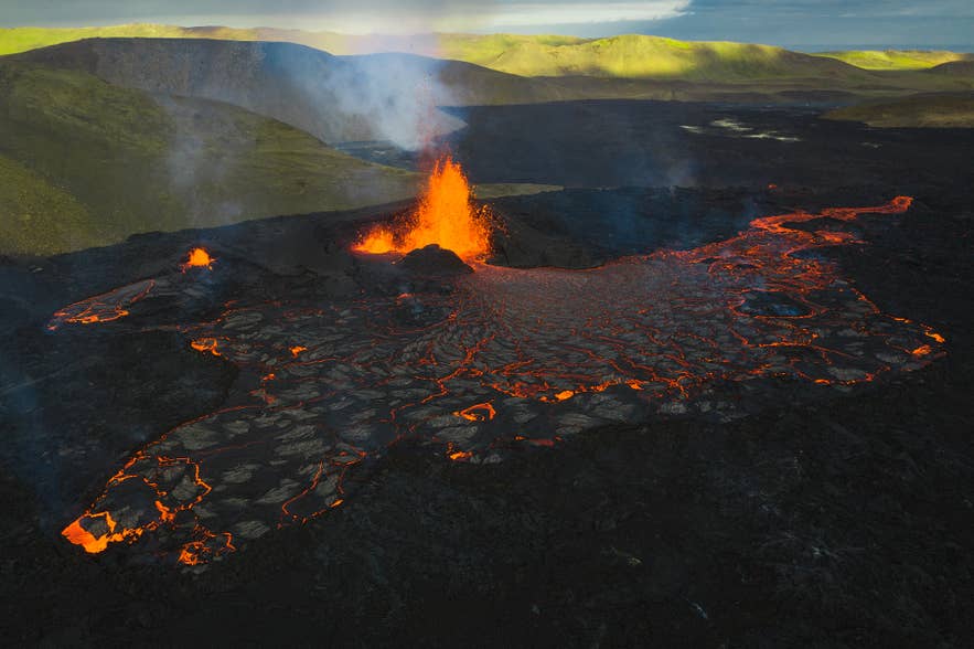 Eruption at Litli-Hrútur volcano in Iceland, with glowing lava flows and smoke against a backdrop of green hills.