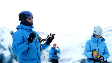 A glacier guide in a blue jacket holds crampons while preparing for a Solheimajokull ice hike in winter conditions.