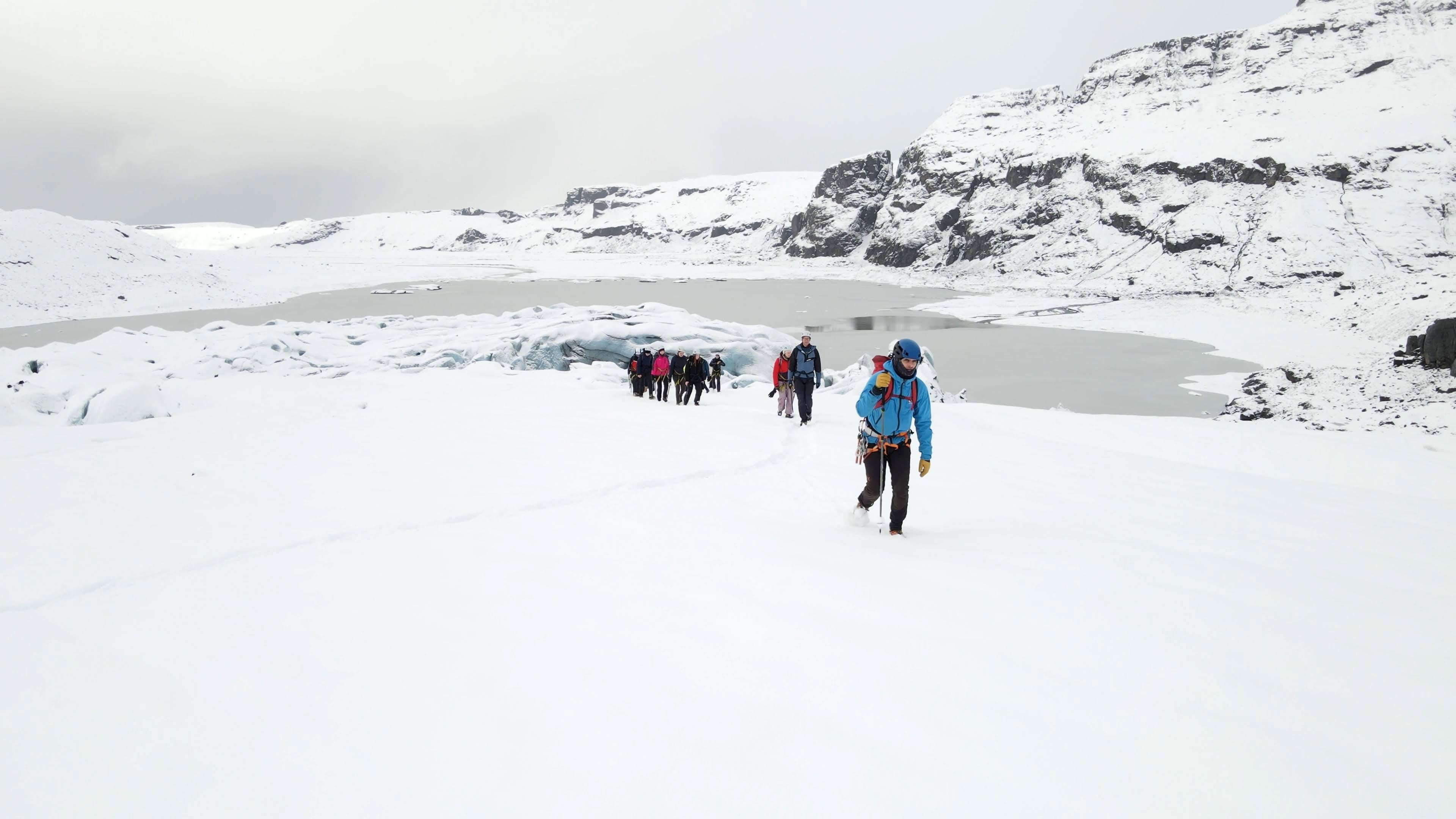 Glacier hikers ascend a snowy slope with the frozen lake and icy cliffs of Solheimajokull Glacier behind them